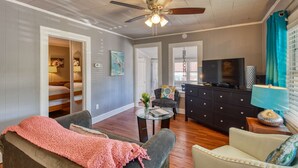 A spacious living room with a ceiling fan, a television, a dark wooden dresser, and a cozy seating area with a pink blanket. - Seaside Cottage (St. Augustine)