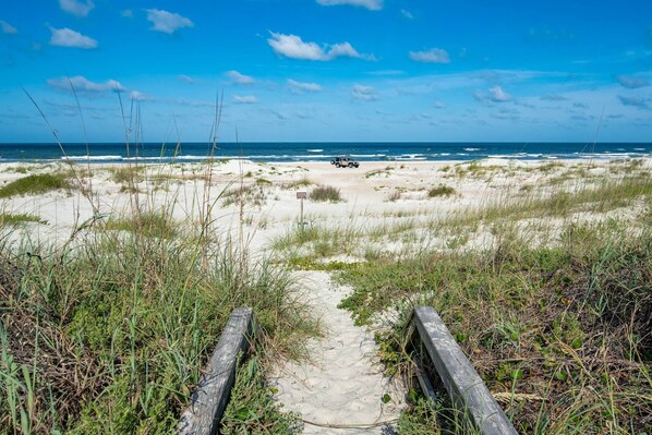Walk along the beach pathway to the ocean, flanked by grassy dunes and a fence.