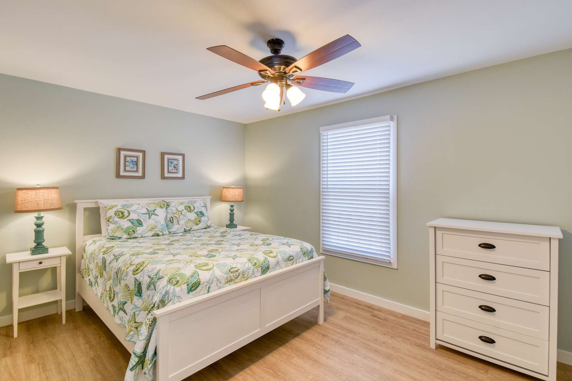 Quarto com cama, duas mesas de cabeceira, cômoda, ventilador de teto e janela com persianas.