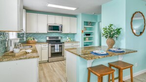 Una cocina con armarios blancos, microondas, horno, encimeras de granito y una pequeña isla con taburetes.