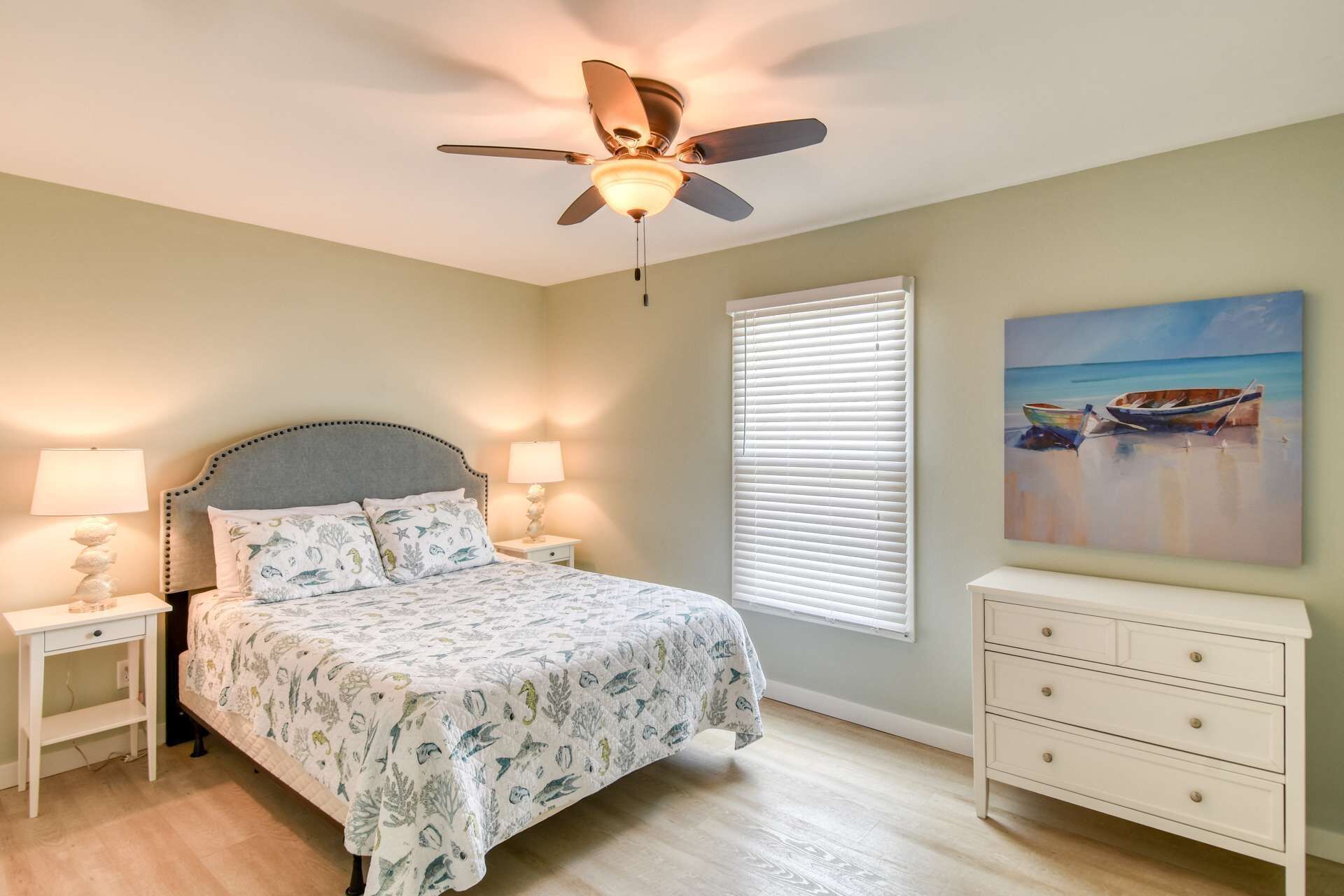 A bedroom with a bed, nightstands, a ceiling fan, a window with blinds, a painting, and a dresser.