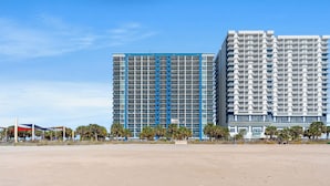 A modern high-rise building with a grid of windows and balconies, situated next to a sandy area with palm trees. - Bay View Resort by Elliott Beach Rentals (Myrtle Beach)