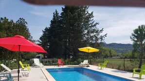A rectangular swimming pool with a tiled edge, surrounded by a concrete deck. There are several lounge chairs and two umbrellas providing shade. The background features a natural landscape with trees and hills. - 2 Bedroom Apartment in Anduze (Anduze)