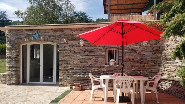 A stone building with a patio area featuring a table and chairs, a red umbrella, and a decorative butterfly on the wall. - 2 Bedroom Apartment in Anduze (Anduze)