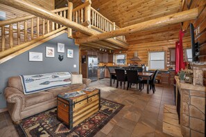 A log cabin interior with a wooden staircase, a dining area, kitchen, and a living room with a sofa and a decorative chest.