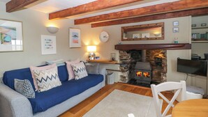 A cozy living room with a blue sofa, a stone fireplace, wooden beams, and a television. - 4 Trenhaile Terrace (Truro)