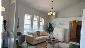 A spacious living room with a beige sofa, a green armchair, a round coffee table, and a large window providing natural light. - Cozy 3-Bedroom Home in North Port (North Port)