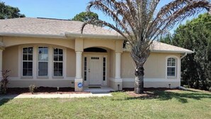 A single-story house with a beige facade, a front porch, and a palm tree in the yard. - Cozy 3-Bedroom Home in North Port (North Port)