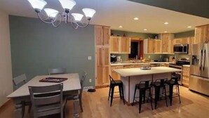 A modern kitchen with wooden cabinets, a central island with bar stools, and a dining area with a table and chairs. - The Good Place: Lakehouse Near Dunbar ATV Trails/On Blue Ox Trails (Fence)