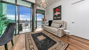 A spacious living room with a large window, a chandelier, a sofa, a chair, and a view of the city. - Penthouse Glory - Sebel (Auckland)