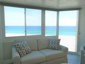 A room with a beige sofa and patterned cushions, large windows with a view of a beach and ocean.