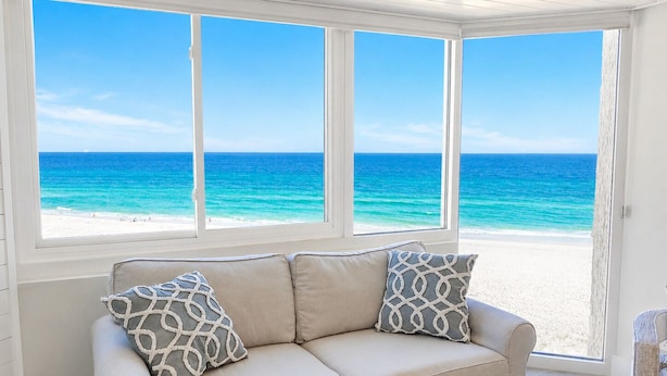 Habitación con un sofá color crema, dos almohadas estampadas y vistas a una playa y al océano a través de amplias ventanas.