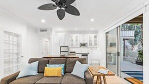 A modern living room with a brown sectional sofa, a ceiling fan, and a view of a kitchen and outdoor area through glass doors. - Sacred Cove Oasis - Indian Wells Tennis and Festival Hideaway (Indian Wells)