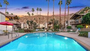 A swimming pool with lounge chairs and umbrellas, surrounded by buildings with balconies and palm trees, set against a mountainous backdrop. - Sacred Cove Oasis - Indian Wells Tennis and Festival Hideaway (Indian Wells)