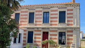 Un bâtiment à deux étages doté d’une façade symétrique, avec une entrée centrale marquée par une porte rouge, de part et d’autre de laquelle se trouvent des fenêtres avec des volets.