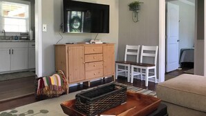 Un salon comprenant un téléviseur, un meuble en bois, deux chaises blanches, une petite table et un panier coloré en fibres tressées.
