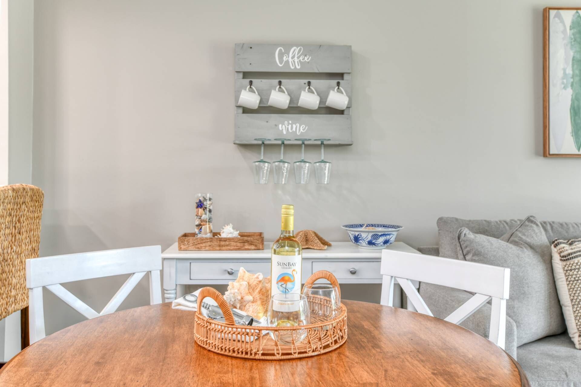 A dining area with a wooden table, wicker chairs, a wicker basket with a bottle, and a wall-mounted wine and coffee rack.
