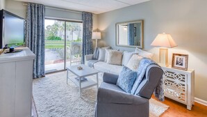 A living room with a grey sofa, white coffee table, and a TV on a stand. There is a window with curtains, a lamp, and a framed picture on a side table. - Ocean and Racquet #3116 (St. Augustine)