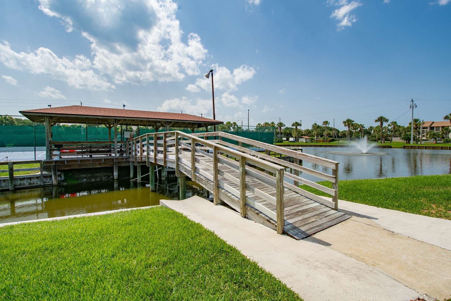 A wooden bridge leading to a covered pavilion over a body of water, with a fountain in the background.