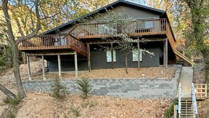 A cabin with a deck overlooking a body of water, surrounded by trees. - Modern lakeside private home, dock, main channel views, off Horsehoe Bend! (Lake Ozark)