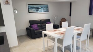 Een moderne woonkamer met een zwarte leren bank, een witte eettafel met vier stoelen, een groot schilderij in een lijst aan de muur en een staande ventilator.