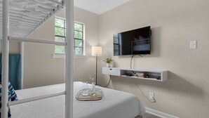 A bedroom with a bunk bed, a mounted TV, a bedside table with a lamp, and a window with a view of trees. - Salty Siesta: Cozy Condo-Walk to Beach-Pools-Golf! (Miramar Beach)