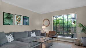 A living room with a grey sofa, a coffee table, a chair, and a large window with a view of greenery. - Salty Siesta: Cozy Condo-Walk to Beach-Pools-Golf! (Miramar Beach)