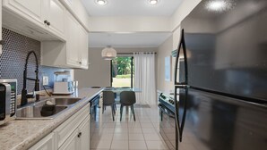 A modern kitchen with white cabinets, a black refrigerator, and a dining area with a table and chairs. - Salty Siesta: Cozy Condo-Walk to Beach-Pools-Golf! (Miramar Beach)