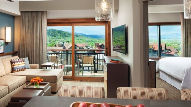 Ein Hotelzimmer mit einem großen Fenster und einem herrlichen Panoramablick auf eine gebirgige Landschaft. Es gibt ein Sofa, einen Esstisch mit Obstschale und einen an der Wand befestigten Fernseher.