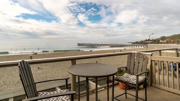 A balcony with a table and chairs, overlooking a beach and a pier.
