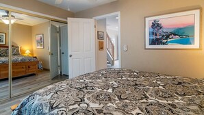A bedroom with a patterned bedspread, a ceiling fan, a door leading to another room, and a framed landscape painting on the wall.