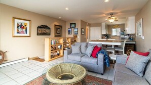 A living room with a grey sofa, a wicker chair, a bookshelf, and a dining area with a white table and chairs.