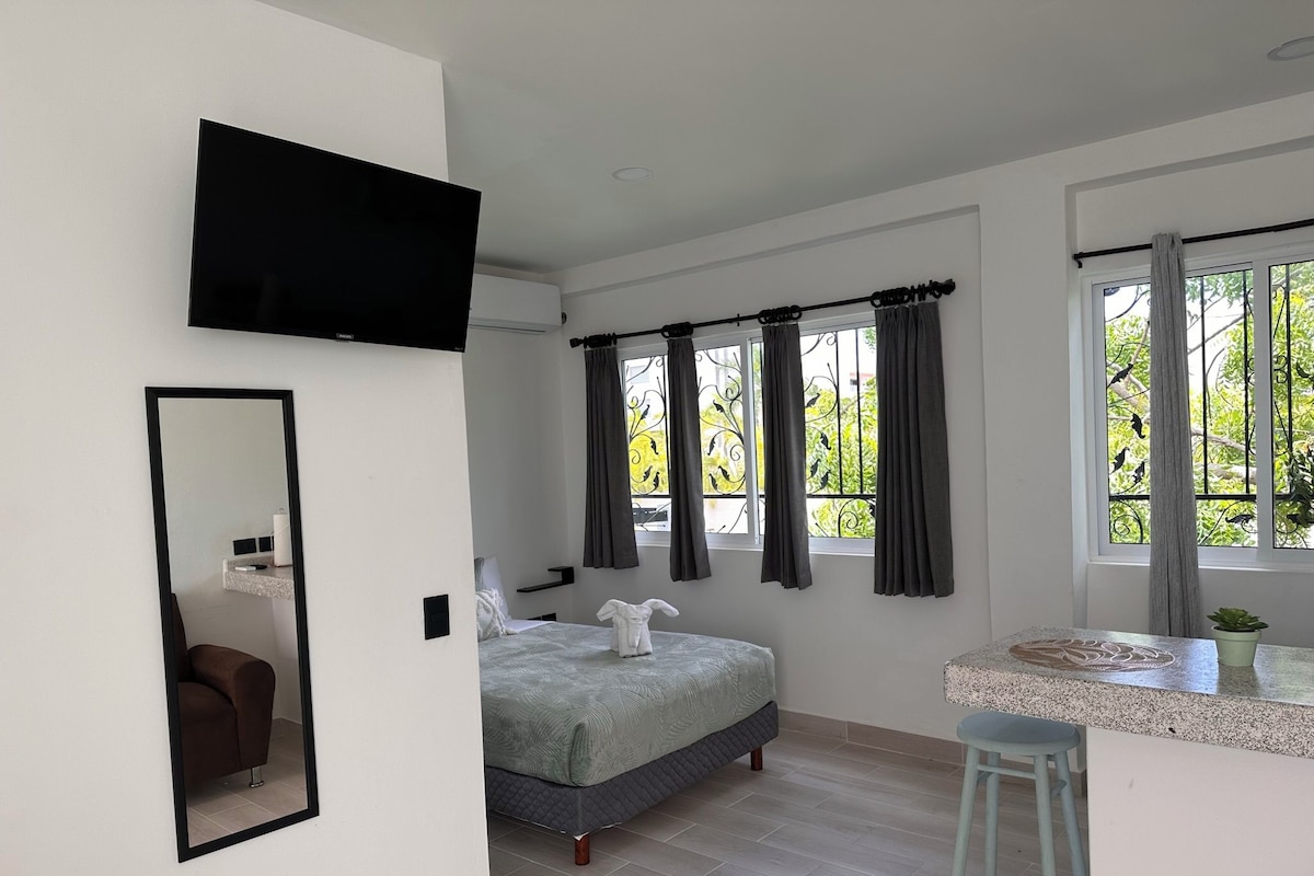 Un dormitorio moderno con cama, televisor montado en la pared, baño con lavamanos y ventana con cortinas.