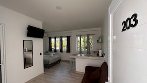 Una habitación moderna y luminosa con una cama, un sofá marrón, una pequeña cocina y un televisor montado en la pared.