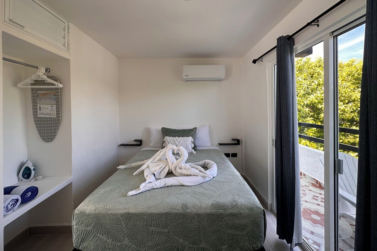 Ein Schlafzimmer mit Bett, Kleiderstange, Bügelbrett und Blick auf Grün durch das Fenster.