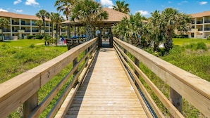 A wooden boardwalk leading to a covered area with palm trees and a building in the background. - Ocean Villas #76 (St. Augustine)