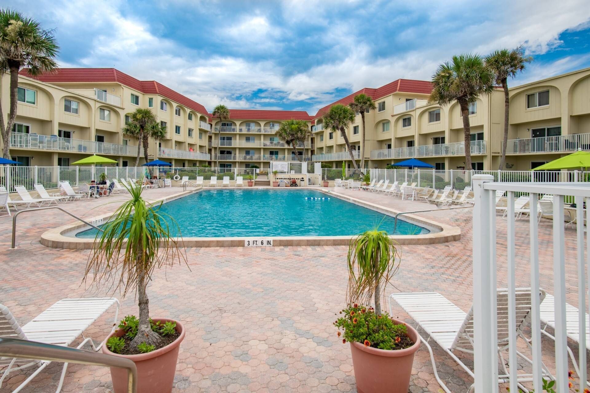 A swimming pool surrounded by lounge chairs and potted plants, with a multi-story building in the background.