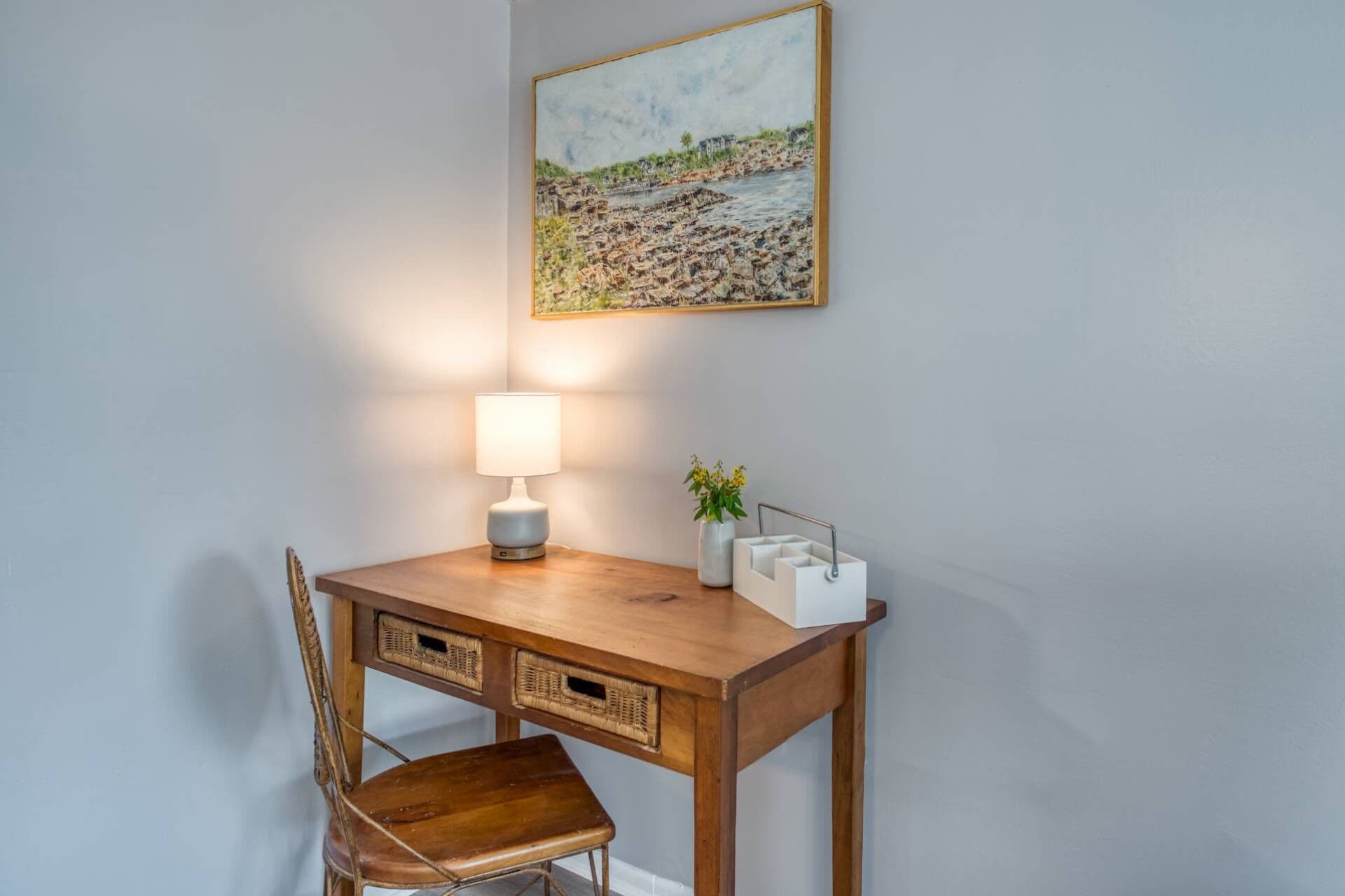 Een houten bureau met een lamp en een vaas met bloemen, een stoel en een schilderij aan de muur.
