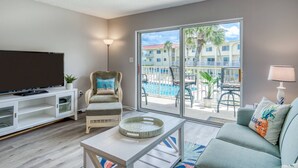 Un salon avec un téléviseur, un fauteuil avec un coussin, une table basse et un canapé. Il y a un balcon avec une piscine et des bâtiments en arrière-plan.
