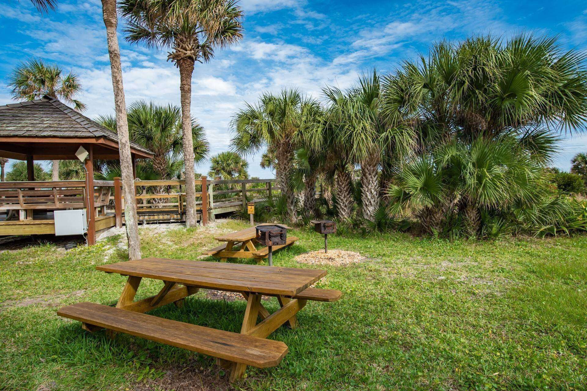 A wooden picnic table with benches, a gazebo, and palm trees in the background.