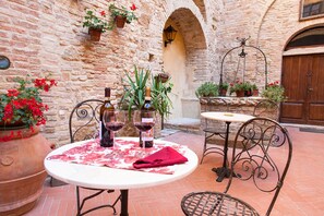 An outdoor patio with a round table, two chairs, and a wine bottle. There are potted plants and a stone wall in the background.