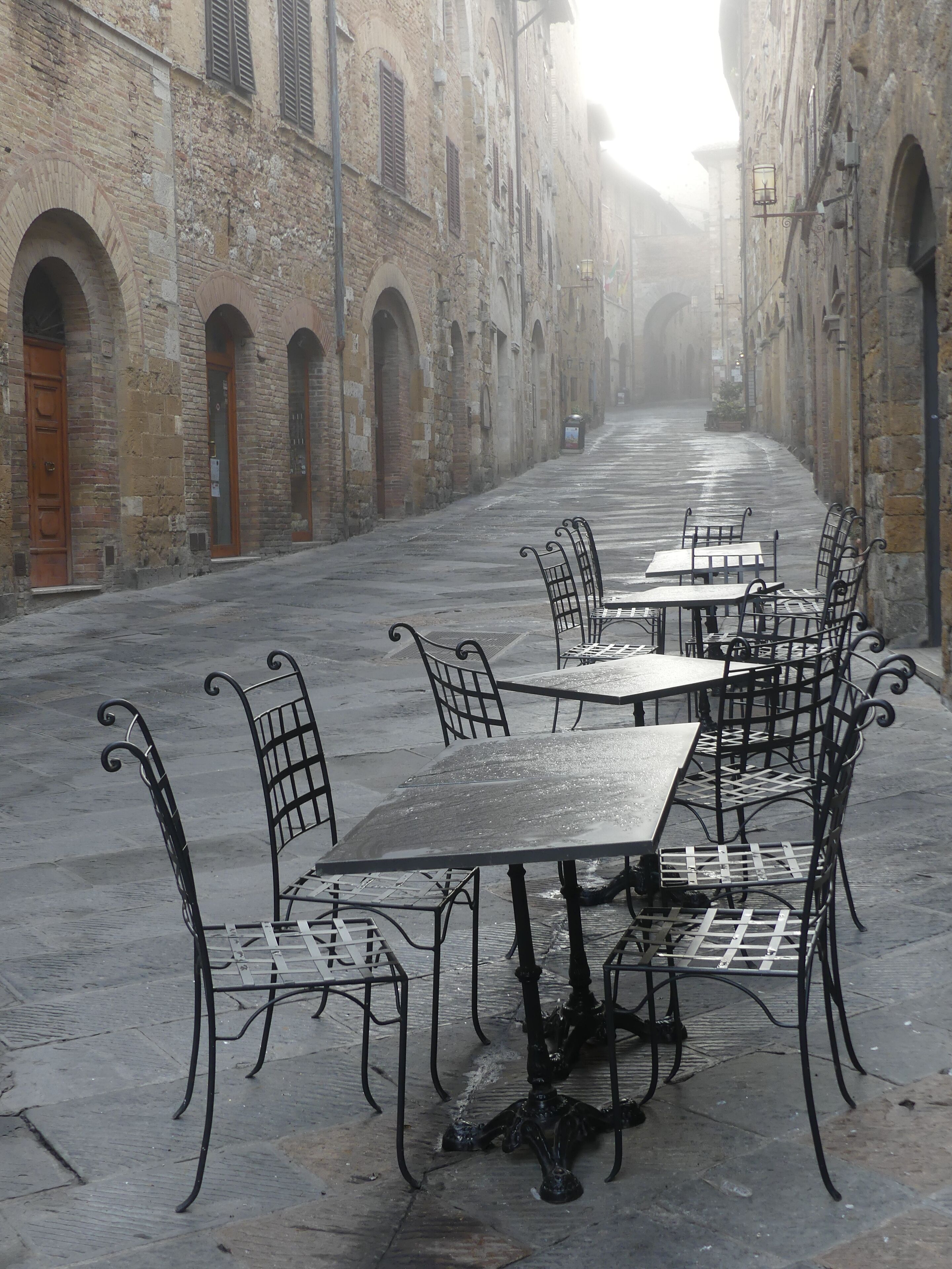 Ein Straßencafé mit Metalltischen und -stühlen vor der Kulisse historischer Steinbauten mit Rundbogenfenstern und -portalen.