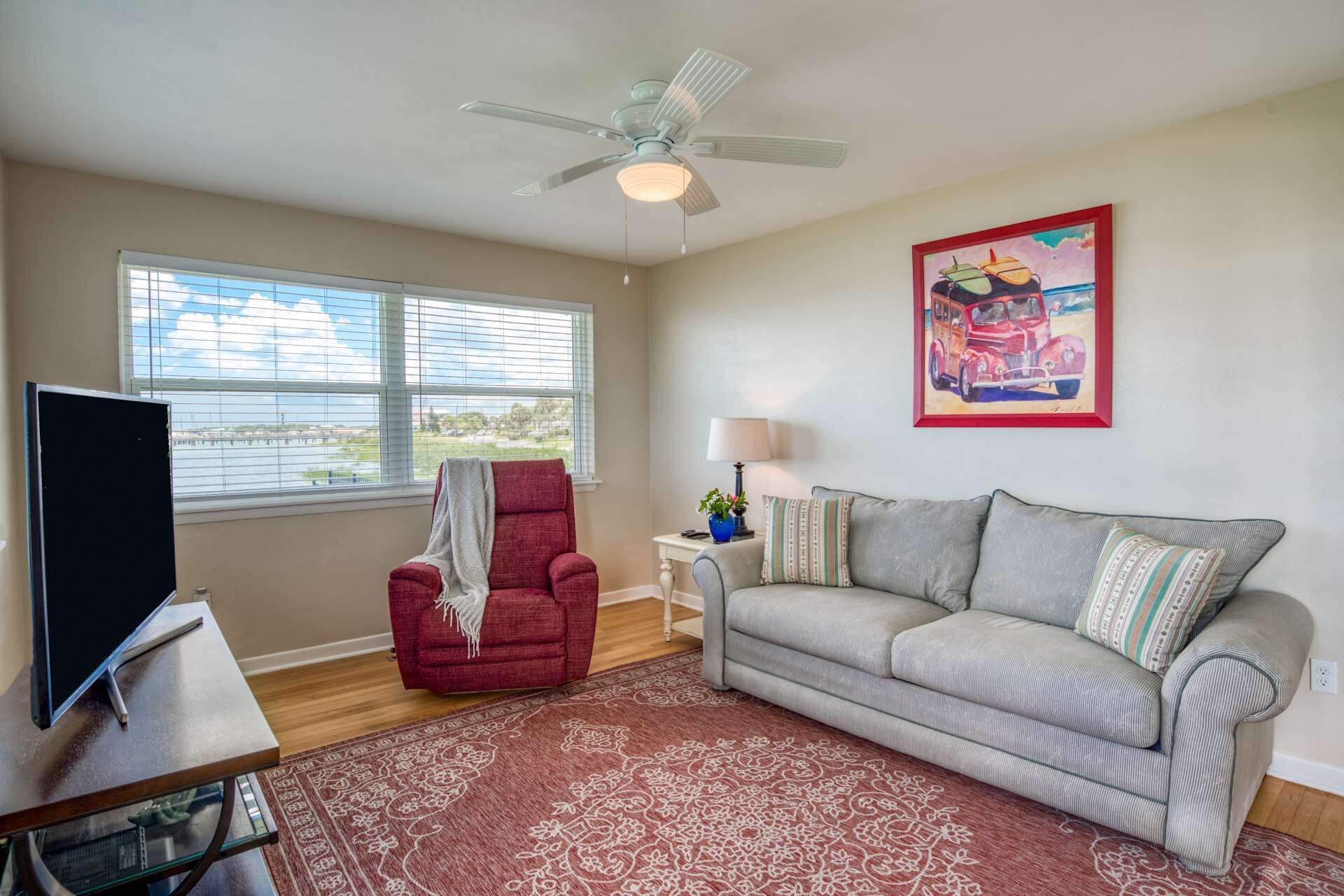 A living room with a red armchair, a grey sofa, a ceiling fan, a painting, and a view through the window.