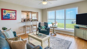 A spacious living room with a large window offering a view of the water, a ceiling fan, a television, and a kitchen area visible through an open door. - Porpoise Point (St. Augustine)
