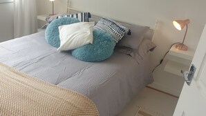 Una cama bien hecha con una colcha a rayas, una almohada blanca y dos almohadas azules y mullidas. Hay una mesita de noche con una lámpara y un enchufe.