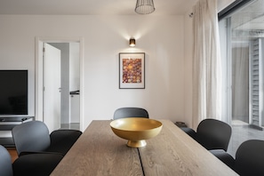 A modern dining area with a wooden table, black chairs, a large gold bowl centerpiece, a framed artwork above the table, and a television in the background.