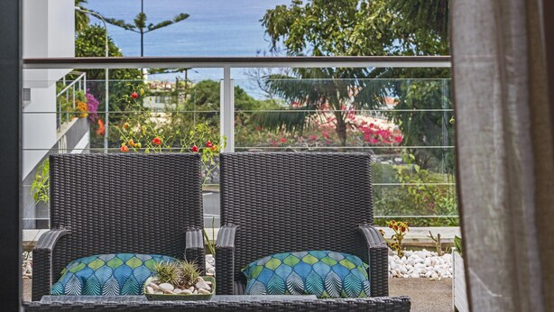 Zwei Korbstühle mit gemusterten Kissen auf einem Balkon mit Blick auf Meer und Garten.