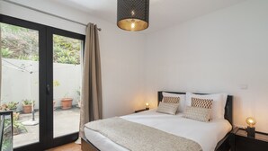 Ein Schlafzimmer mit einem großen Bett, zwei Nachttischen, einer Deckenleuchte und einer Glasschiebetür zu einem Außenbereich mit Blumentöpfen.