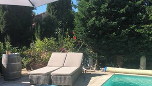 A swimming pool with a lounge area, including a sofa and a chair, and a large umbrella providing shade. - Air-conditioned town house with heated swimming pool (Arles)