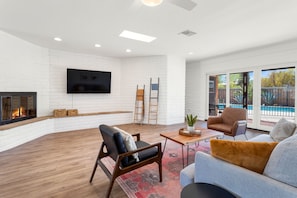 A modern living room with a fireplace, a flat-screen TV, and a view of a pool through large sliding glass doors.